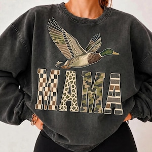 May include: A charcoal grey acid-wash crewneck sweatshirt featuring a flying mallard duck graphic above the word MAMA. The letters are filled with distinct patterns: a brown checkerboard, leopard print, green camouflage, and horizontal stripes.