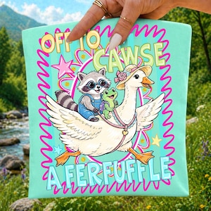 May include: A light teal tote bag with a whimsical illustration. A raccoon wearing glasses and overalls, and a green frog ride on a goose wearing a hat. The text reads "Off to Cawse A Ferfuffle" in a playful font, surrounded by pink and yellow accents.