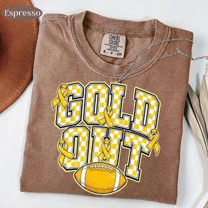 Puede incluir: Una camiseta marr&oacute;n de manga corta con cuello redondo y etiqueta Comfort Colors. El gr&aacute;fico presenta el texto GOLD OUT en un patr&oacute;n de cuadros amarillos y blancos, acentuado con cintas amarillas de concienciaci&oacute;n y un bal&oacute;n de f&uacute;tbol.