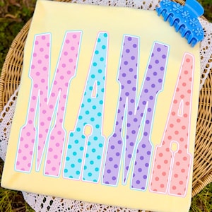 May include: A light yellow t-shirt with the word "MAMA" in large, colorful letters. The letters are outlined in white and filled with polka dots in pink, blue, purple, and peach. A blue hair clip with flower details rests on the top right corner.