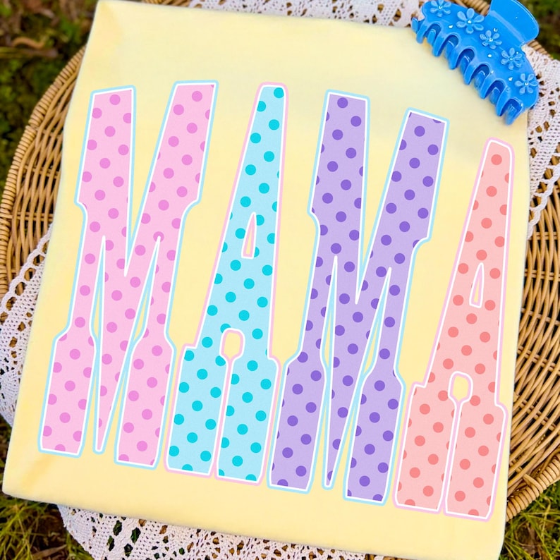 May include: A light yellow t-shirt with the word "MAMA" in large, colorful letters. The letters are outlined in white and filled with polka dots in pink, blue, purple, and peach. A blue hair clip with flower details rests on the top right corner.