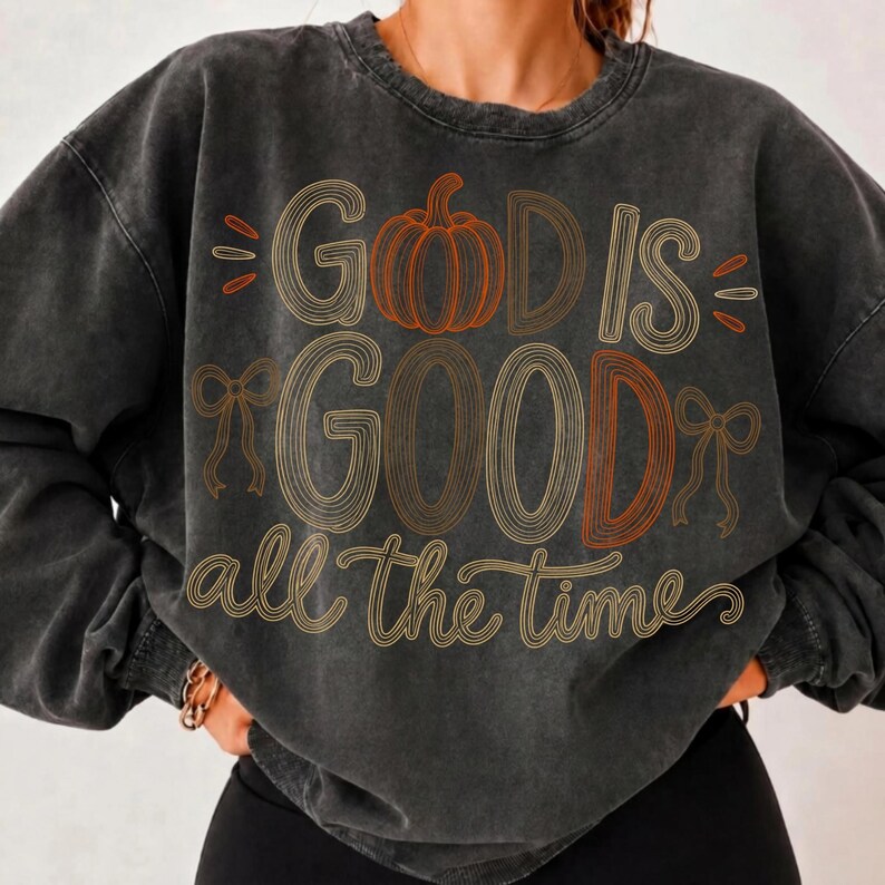 May include: A charcoal grey mineral-washed crewneck sweatshirt featuring a graphic print in orange and cream tones. The design includes a pumpkin illustration, two small bows, and the text "GOD IS GOOD all the time" in a stylized, layered font.