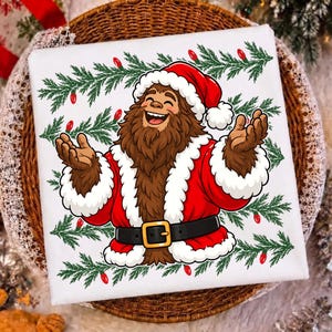 Puede incluir: Una camiseta blanca con el gr&aacute;fico de un Bigfoot marr&oacute;n vestido con un traje de Pap&aacute; Noel rojo con ribetes de piel blanca, un cintur&oacute;n negro con hebilla dorada y un gorro a juego, rodeado de ramas de pino verdes y luces navide&ntilde;as rojas.