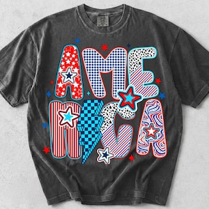 May include: A charcoal gray short-sleeve t-shirt featuring the word AMERICA in large, stylized letters. Each letter displays a different pattern, including red stars, blue and white checkers, red and white stripes, and blue and white swirls.