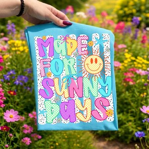 May include: A light blue t-shirt featuring a colorful graphic with the text "MADE FOR SUNNY DAYS" in a playful font. The design includes a yellow checkered smiley face sun, small flowers, starfish, and blue bows, all set against a background of black dots.