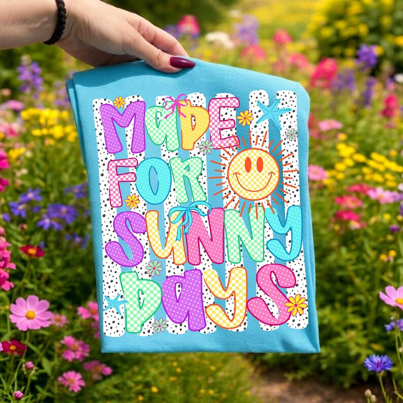 May include: A light blue t-shirt featuring a colorful graphic with the text "MADE FOR SUNNY DAYS" in a playful font. The design includes a yellow checkered smiley face sun, small flowers, starfish, and blue bows, all set against a background of black dots.