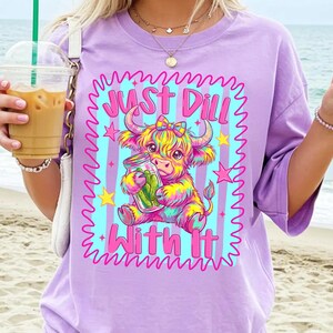 Puede incluir: Una camiseta de color morado claro con un gr&aacute;fico colorido de una vaca highland sosteniendo un frasco de pepinillos. El dise&ntilde;o incluye un fondo de rayas azul claro, estrellas rosas y el texto "JUST DILL WITH IT" en una fuente rosa.