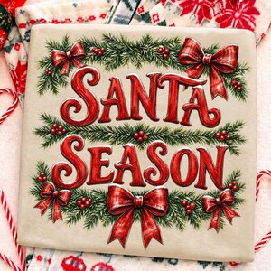 以下が含まれることがあります： ベージュのTシャツに、赤い文字で「SANTA SEASON」とスタイリッシュなフォントでプリントされています。デザインは緑の松の枝、赤いベリー、赤のチェック柄のリボンで縁取られています。グラフィックは生地の中央に配置され、祝祭的な雰囲気を醸し出しています。