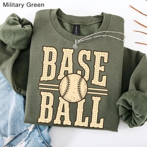 Puede incluir: Una sudadera de cuello redondo color verde militar con un gran estampado gr&aacute;fico que muestra el texto BASE BALL en una fuente beige punteada. Una ilustraci&oacute;n central de una pelota de b&eacute;isbol se sit&uacute;a entre las dos palabras. Un collar de plata descansa en el cuello.