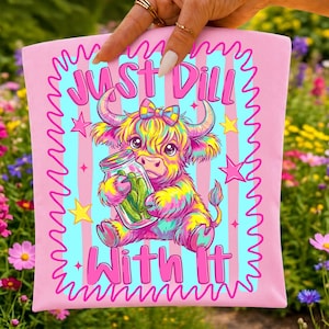 Puede incluir: Un cuadrado de tela rosa claro con una ilustraci&oacute;n colorida de una vaca highland sosteniendo un frasco de vidrio con pepinillos. El texto "JUST DILL WITH IT" est&aacute; impreso en letras rosas en negrita. El dise&ntilde;o incluye rayas rosas, estrellas y un borde ondulado.