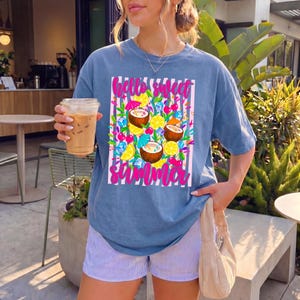 May include: A blue short-sleeve t-shirt featuring a graphic print of coconuts, lemon slices, cherries, and ice cubes over a pink and white striped background with the text "hello sweet summer" in bright pink script lettering.