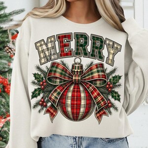 Puede incluir: Una sudadera de color crema con la palabra MERRY en una fuente de cuadros sobre un gr&aacute;fico de un adorno navide&ntilde;o. El adorno est&aacute; decorado con un gran lazo de cuadros, ramas de pino verdes y bayas rojas, creando un dise&ntilde;o festivo.