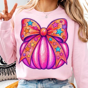 May include: A light pink sweatshirt featuring a large, vibrant graphic of a pumpkin decorated with a prominent bow. The design is rendered in a scribbled, sketch-like style with bright orange, magenta, and purple hues, accented by blue and yellow stars.