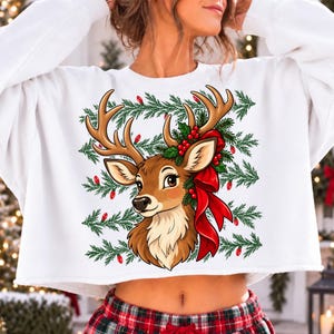 May include: A white cropped sweatshirt featuring a graphic of a brown deer head with large antlers, adorned with a red bow, green pine branches, and red berries. The background of the graphic includes a pattern of green pine needles and red lights.