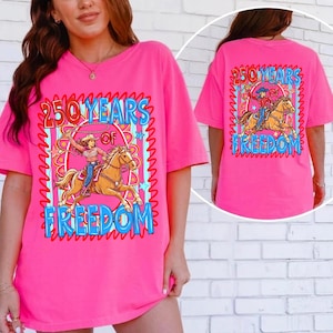 Puede incluir: Una camiseta rosa brillante con un estampado gr&aacute;fico de una vaquera montando un caballo marr&oacute;n mientras sostiene un lazo. El texto 250 YEARS OF FREEDOM est&aacute; impreso en azul con un borde rojo. El dise&ntilde;o incluye un marco circular y estrellas.