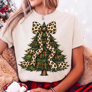 May include: A cream-colored short-sleeve t-shirt featuring a graphic of a green Christmas tree with a red and green plaid pattern. The tree is adorned with a large, flowing ribbon featuring a tan and black leopard print pattern.