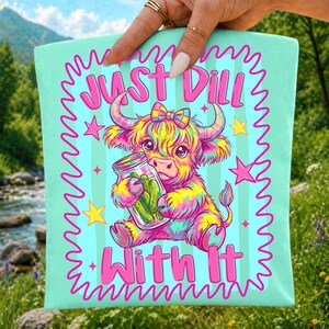 Puede incluir: Un art&iacute;culo de tela verde menta claro con una ilustraci&oacute;n colorida de una vaca highland esponjosa con un lazo, sosteniendo un frasco de vidrio con pepinillos. El texto Just Dill With It aparece en letras rosa brillante, rodeado de estrellas.
