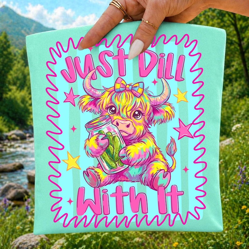 Puede incluir: Un art&iacute;culo de tela verde menta claro con una ilustraci&oacute;n colorida de una vaca highland esponjosa con un lazo, sosteniendo un frasco de vidrio con pepinillos. El texto Just Dill With It aparece en letras rosa brillante, rodeado de estrellas.