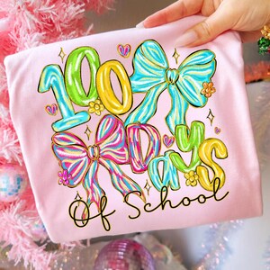 Op de afbeelding: Lichtroze T-shirt met de tekst "100 Days of School" in kleurrijke, speelse letters. Het ontwerp bevat grote strikken, harten en bloemen in levendige tinten blauw, roze, geel en groen.