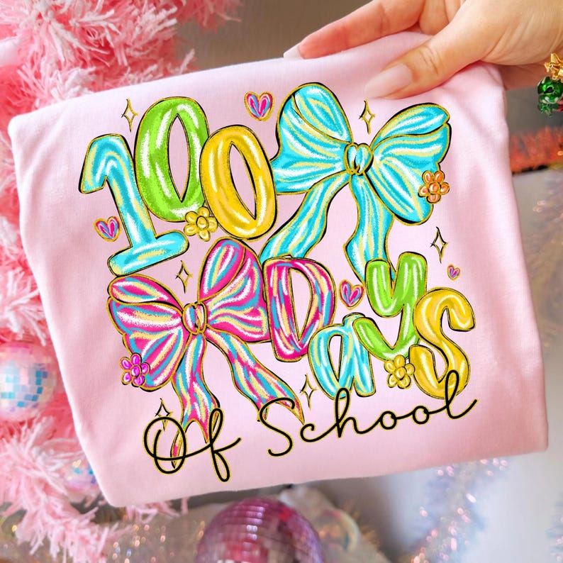 Op de afbeelding: Lichtroze T-shirt met de tekst "100 Days of School" in kleurrijke, speelse letters. Het ontwerp bevat grote strikken, harten en bloemen in levendige tinten blauw, roze, geel en groen.