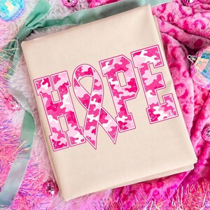 Peut inclure: Un t-shirt beige avec le mot HOPE imprim&eacute; en grandes lettres majuscules. La lettre O est remplac&eacute;e par un ruban de sensibilisation rose. Les lettres pr&eacute;sentent un motif camouflage rose et blanc avec un fin contour rose autour de chaque caract&egrave;re.