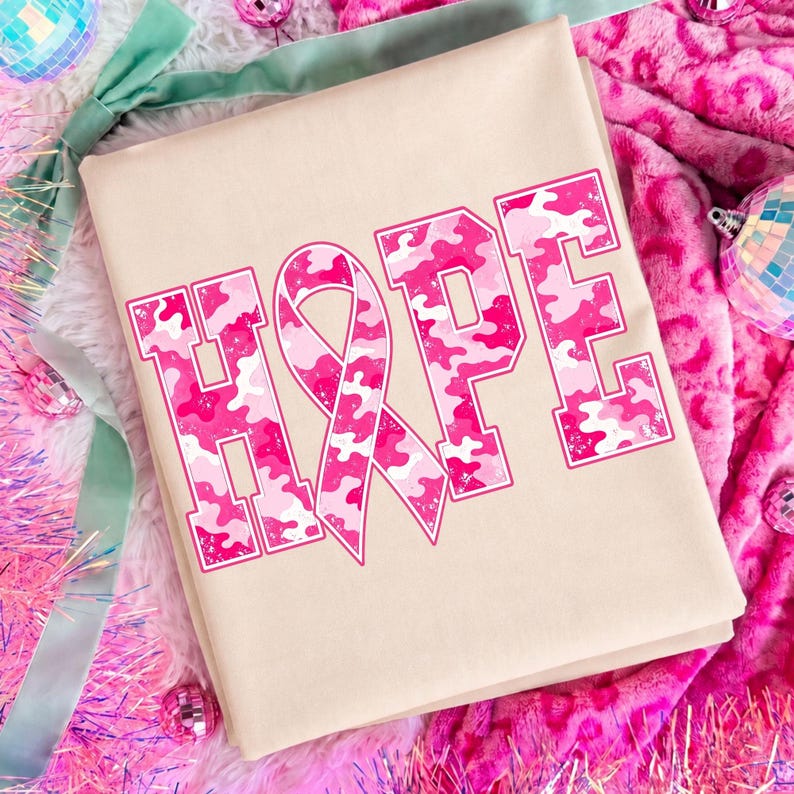 Peut inclure: Un t-shirt beige avec le mot HOPE imprim&eacute; en grandes lettres majuscules. La lettre O est remplac&eacute;e par un ruban de sensibilisation rose. Les lettres pr&eacute;sentent un motif camouflage rose et blanc avec un fin contour rose autour de chaque caract&egrave;re.