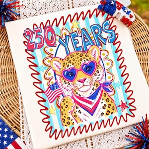 May include: A white t-shirt featuring a graphic of a leopard wearing heart-shaped sunglasses and a patriotic bandana, holding an American flag. The text 250 YEARS is printed above in blue and pink. The design includes red wavy borders and stars.