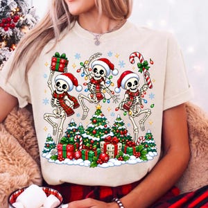 Op de afbeelding: Een beige t-shirt met een afbeelding van drie skeletten met kerstmutsen en sjaals, dansend rond kerstbomen en cadeautjes. De skeletten zijn versierd met kleurrijke kerstverlichting en het ontwerp bevat diverse sneeuwvlokken.