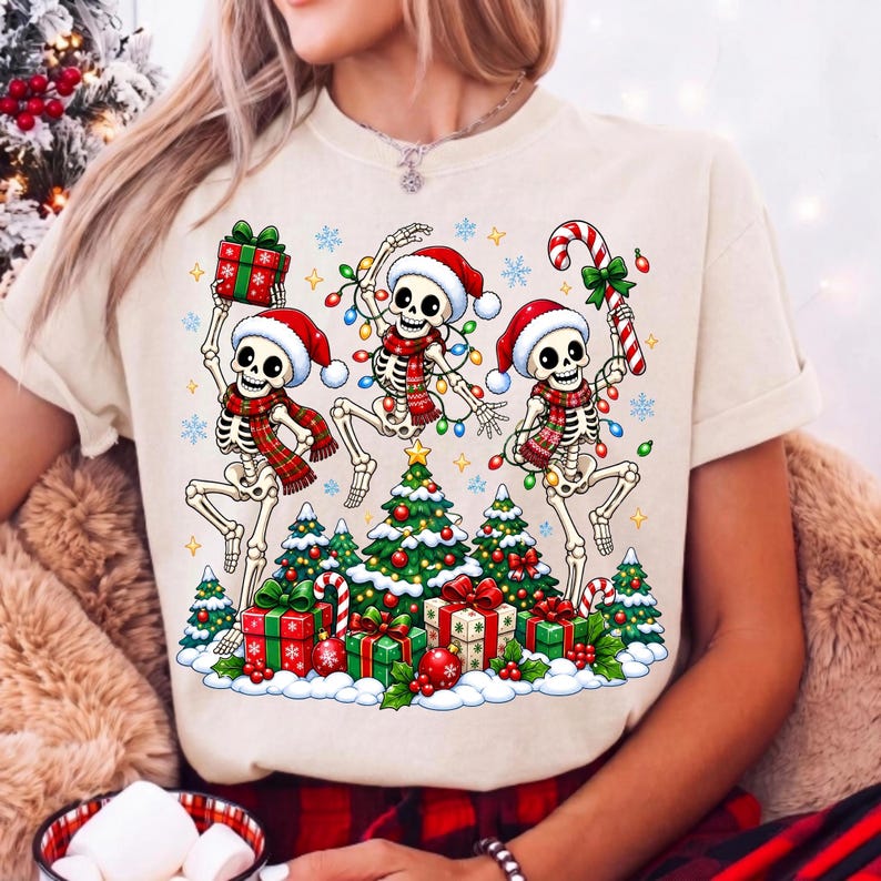 Op de afbeelding: Een beige t-shirt met een afbeelding van drie skeletten met kerstmutsen en sjaals, dansend rond kerstbomen en cadeautjes. De skeletten zijn versierd met kleurrijke kerstverlichting en het ontwerp bevat diverse sneeuwvlokken.