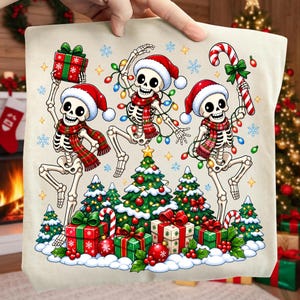 Op de afbeelding: Een beige draagtas met een grafische print van drie skeletten met kerstmutsen en geruite sjaals, dansend rond kerstbomen en cadeaus. De skeletten houden een rood-groen cadeau, een lichtsnoer en een zuurstok vast.