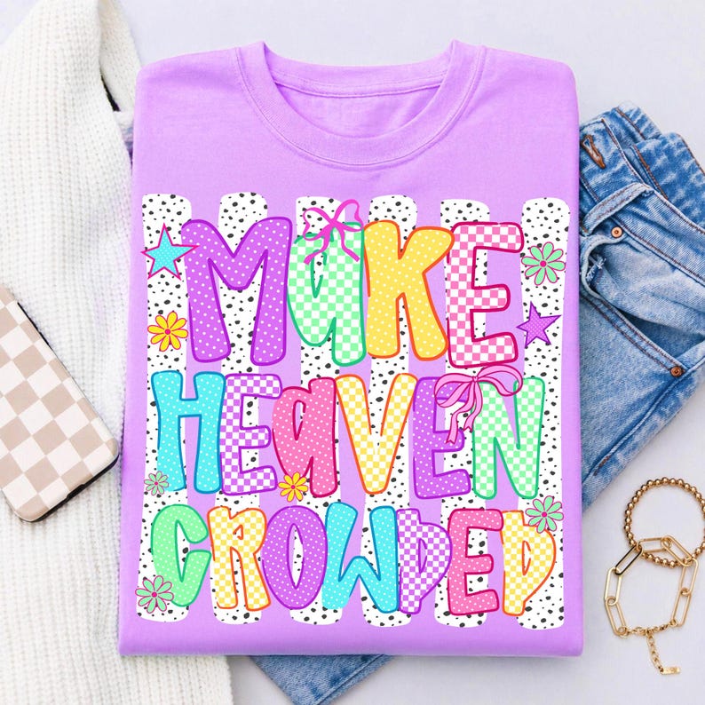 May include: A light purple t-shirt featuring the text MAKE HEAVEN CROWDED in colorful, patterned block letters. The design includes pink bows, small stars, and flowers over vertical white stripes with black dots, creating a vibrant graphic.
