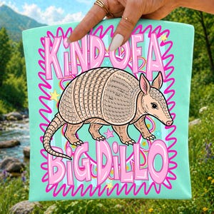 Pu&ograve; includere: Una borsa tote verde menta chiaro con un'illustrazione centrale di un armadillo color marrone chiaro. Il testo "KIND OF A BIG DILLO" &egrave; stampato in grandi lettere rosa sopra e sotto l'animale, circondato da un bordo rosa ondulato e stelle gialle.
