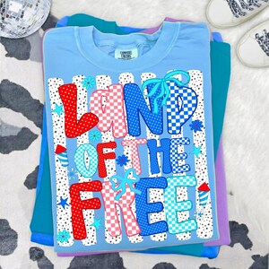 May include: A light blue t-shirt featuring the text LAND OF THE FREE in red, white, and blue checkered and polka dot patterns. The design includes light blue bows, small blue flowers, stars, and firework graphics over vertical black dotted lines.