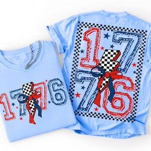 Op de afbeelding: Twee lichtblauwe t-shirts met een afbeelding van het getal 1776 in een rood en blauw schets-lettertype. Een zwart-wit geblokte bliksemschicht staat in het midden, versierd met een rode en blauwe strik, omringd door kleine sterren.