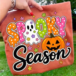May include: A rust-colored fabric item featuring a graphic print with the text "SPOOKY SEASON". The design includes a white ghost, an orange jack-o'-lantern, and colorful polka-dot lettering in orange, pink, and lime green, accented by small flowers.