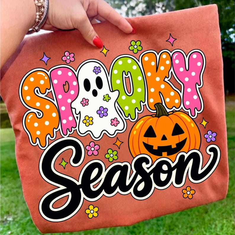 May include: A rust-colored fabric item featuring a graphic print with the text "SPOOKY SEASON". The design includes a white ghost, an orange jack-o'-lantern, and colorful polka-dot lettering in orange, pink, and lime green, accented by small flowers.
