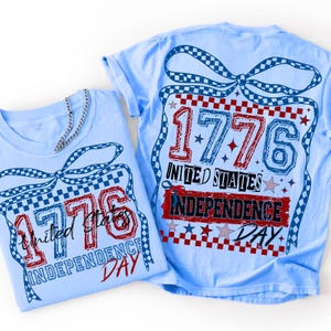 Może przedstawiać: Dwie jasnoniebieskie koszulki z grafiką przedstawiającą kokardę w kratkę nad napisem 1776 UNITED STATES INDEPENDENCE DAY. Projekt zawiera czerwone, białe i niebieskie kolory z akcentami w kształcie gwiazd oraz teksturowany styl czcionki.