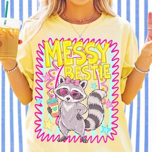 May include: A pale yellow t-shirt featuring a graphic of a raccoon wearing a pink bow and heart-shaped glasses, holding a drink. The text MESSY BESTIE is printed in large yellow letters above the raccoon, surrounded by a pink squiggly border.