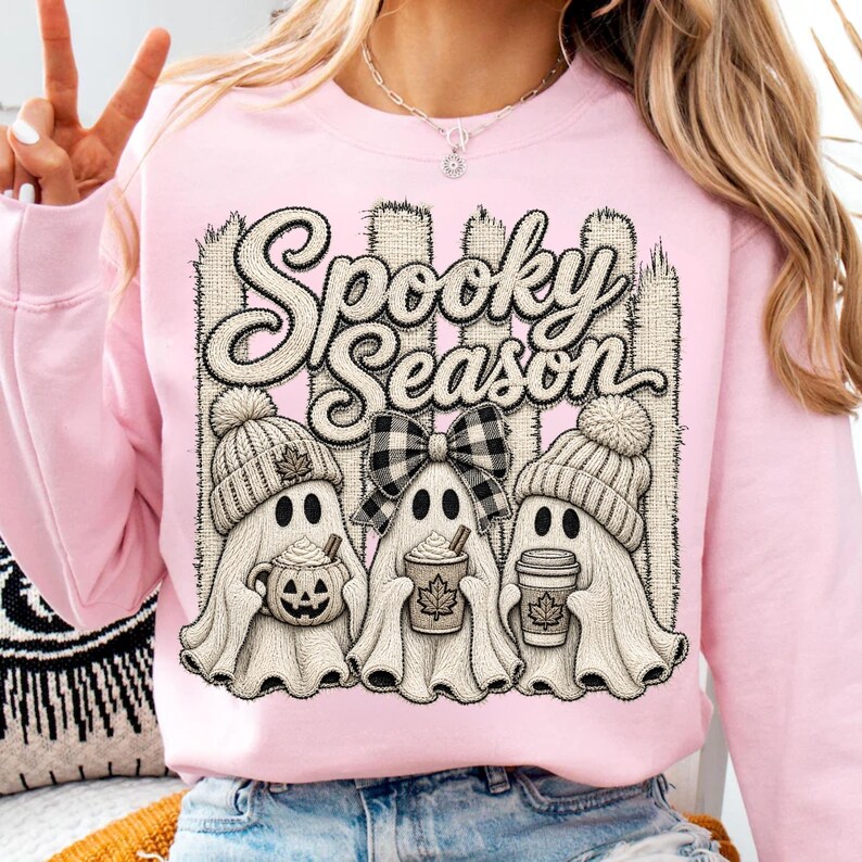 May include: A light pink crewneck sweatshirt featuring a graphic print of three ghosts wearing knit hats. The center ghost wears a black and white plaid bow. The ghosts hold a pumpkin mug and coffee cups. The text reads Spooky Season.