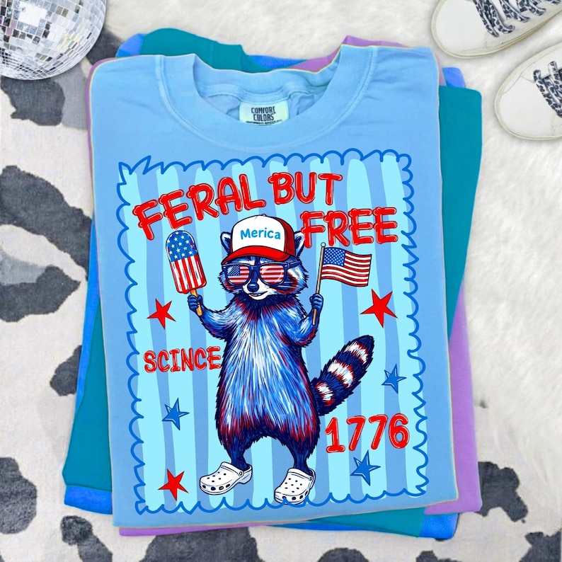 May include: A light blue t-shirt featuring a graphic of a raccoon wearing a red and white hat with the text Merica, red and blue sunglasses, and white clogs. The raccoon holds a flag-patterned popsicle and a US flag. Text reads FERAL BUT FREE 1776.