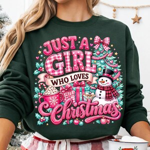 May include: A dark green sweatshirt featuring a graphic print with the text "JUST A GIRL WHO LOVES CHRISTMAS". The design includes a snowman, a decorated Christmas tree, wrapped gifts, and a mug of hot cocoa with a candy cane, all in festive colors.