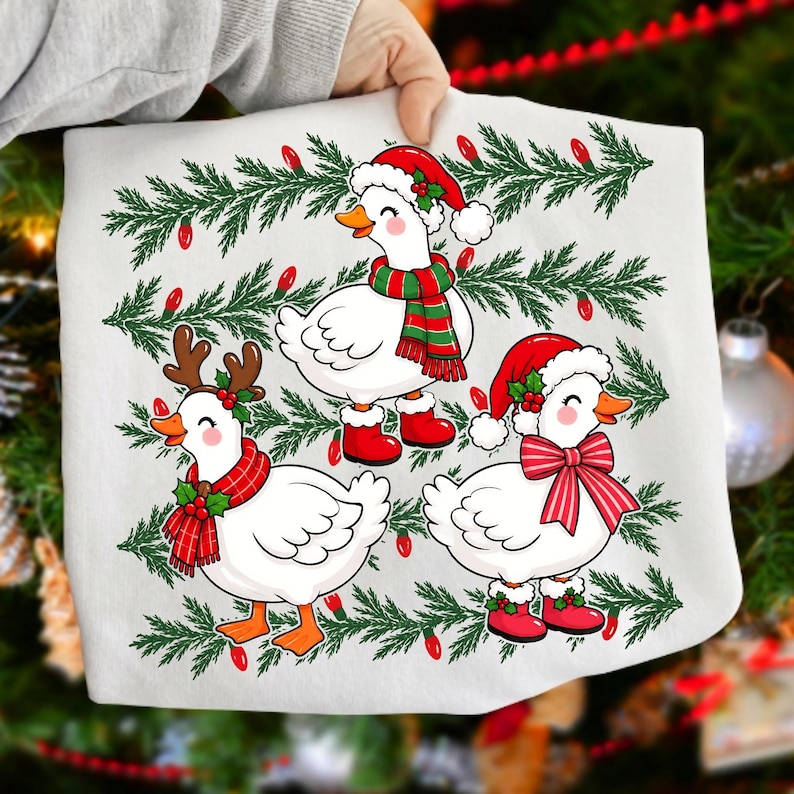 Może przedstawiać: Biały t-shirt z grafiką trzech białych gęsi udekorowanych na święta Bożego Narodzenia. Jedna gęś ma czapkę Mikołaja i szalik, druga poroże renifera i czerwony szalik, a trzecia czapkę Mikołaja i dużą czerwoną kokardę.