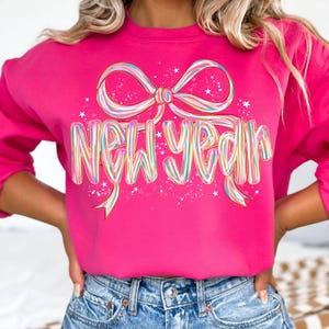 May include: A bright pink long-sleeve sweatshirt featuring a large, colorful graphic of a ribbon bow above the text "NEW YEAR". The lettering and bow design incorporate a multi-colored, iridescent-style striped pattern with small white stars.