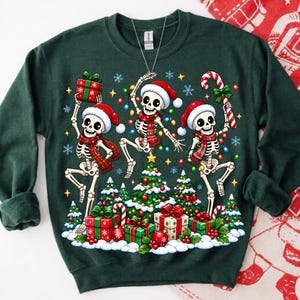 Op de afbeelding: Een donkergroen sweatshirt met lange mouwen en een grafische print van drie dansende skeletten met kerstmutsen en rood geruite sjaals. De skeletten worden omringd door een kerstboom, ingepakte cadeaus, zuurstokken en kleurrijke lichtjes.