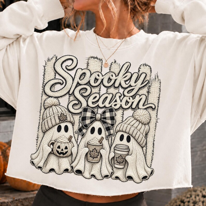 May include: A cream-colored long-sleeve sweatshirt featuring a graphic print with the text Spooky Season. The design includes three ghosts wearing knit hats, one with a buffalo plaid bow, holding a pumpkin mug and two coffee cups.
