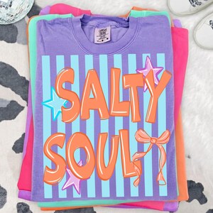 May include: A stack of colorful t-shirts in shades of purple, pink, green, and orange. The top shirt is purple with the words "SALTY SOUL" in orange, outlined in white, over blue vertical stripes. Star and bow graphics are included.