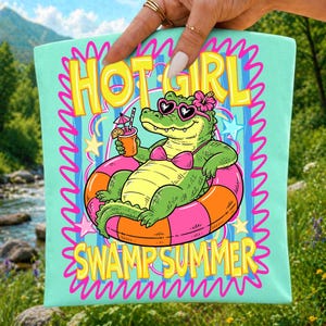 K&ouml;nnte beinhalten: Ein hellminzgr&uuml;ner Stoffartikel mit einer Grafik eines gr&uuml;nen Alligators, der eine herzf&ouml;rmige Sonnenbrille und ein rosa Bikini-Oberteil tr&auml;gt, in einem orange-rosa Schwimmring treibt, ein Getr&auml;nk h&auml;lt und den Text HOT GIRL SWAMP SUMMER zeigt.