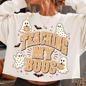 May include: A white long-sleeve sweatshirt featuring a graphic print with the text TEACHING MY BOOS in orange and white gingham. The design includes four white ghosts, two holding orange pumpkins, and small black bats and stars scattered around.