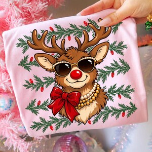 Puede incluir: Una camiseta rosa claro con el gr&aacute;fico de un reno marr&oacute;n que lleva gafas de sol con montura dorada, un collar de perlas de varias vueltas y un gran lazo rojo, rodeado por ramas de pino verdes y peque&ntilde;as luces rojas.