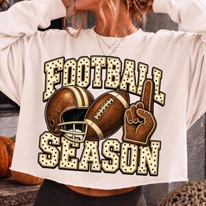 Puede incluir: Una sudadera corta de color crema con un estampado gr&aacute;fico que incluye el texto FOOTBALL SEASON en una fuente de puntos. El dise&ntilde;o presenta un casco de f&uacute;tbol americano marr&oacute;n, un bal&oacute;n de f&uacute;tbol y un dedo de espuma marr&oacute;n.